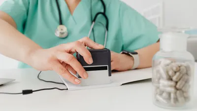 Zdravotník v zeleném oděvu razítkuje dokument, na stole je lahvička s kapslemi a stetoskop